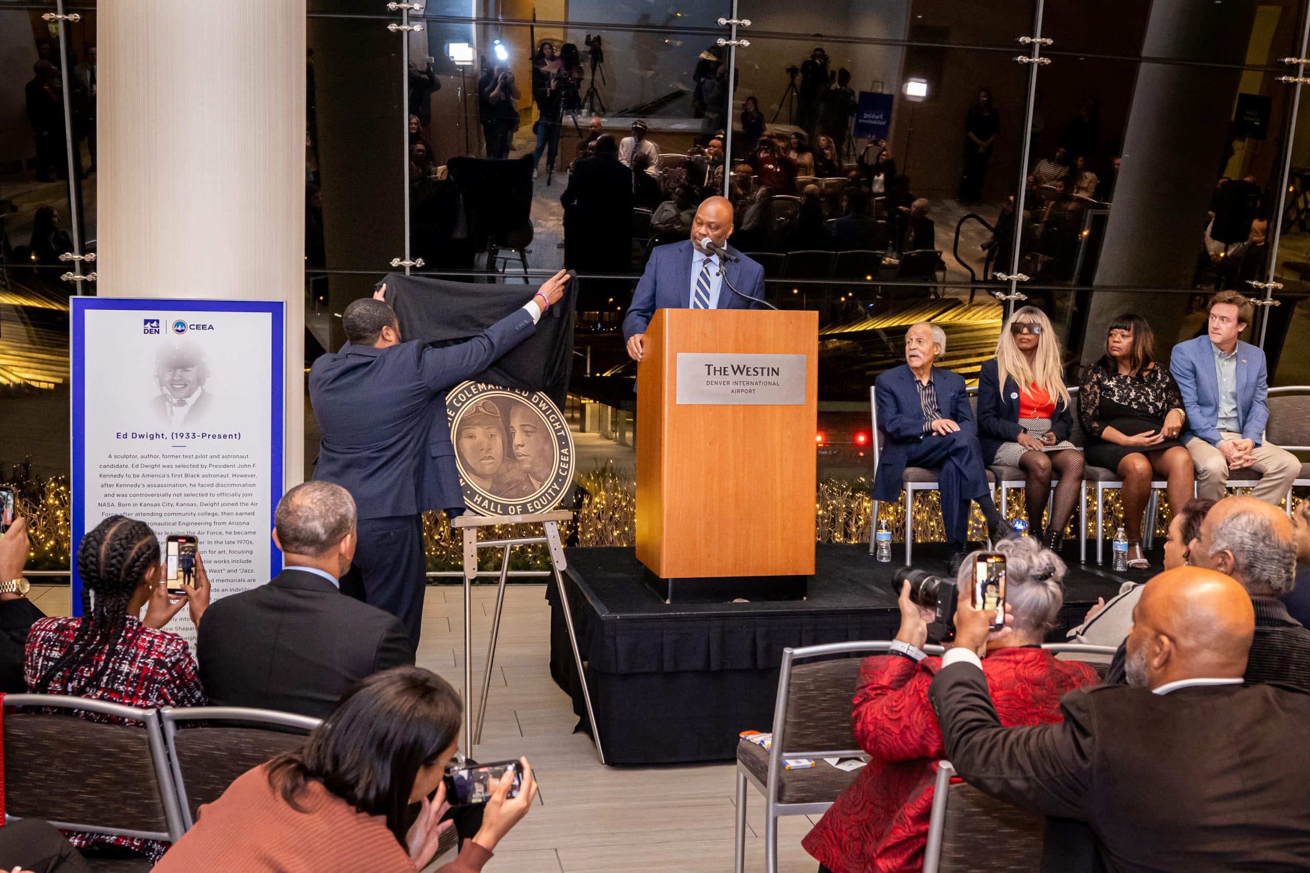 DEN CEO Phil Washington speaking at podium while dedication plaque is unveiled. Next to Phil are Ed Dwight, Jacqueline Withers, Gigi Coleman and Mayor Mike Johnston